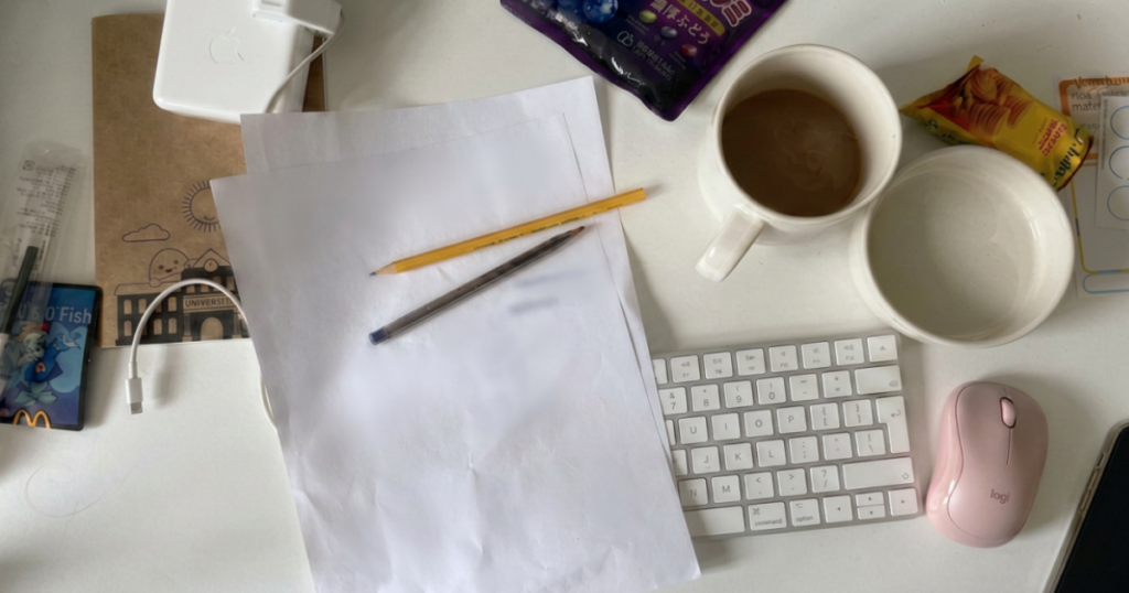 Overhead view of a real messy desk with blank paper, pencil, coffee cups, pink mouse, Apple keyboard and snacks — notes everywhere but nothing useful written down